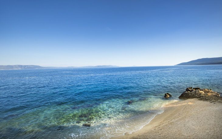 Im Vordergrund ein fein kieseliger Strand und im Hintergrund das blaue Meer und der blaue Himmel